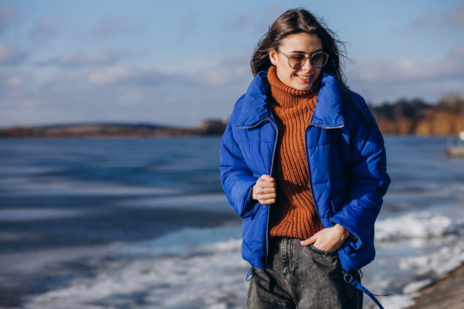 A woman in a cobalt puffer jacket and rust knit sweater on a frozen lakeshore in winter sun.