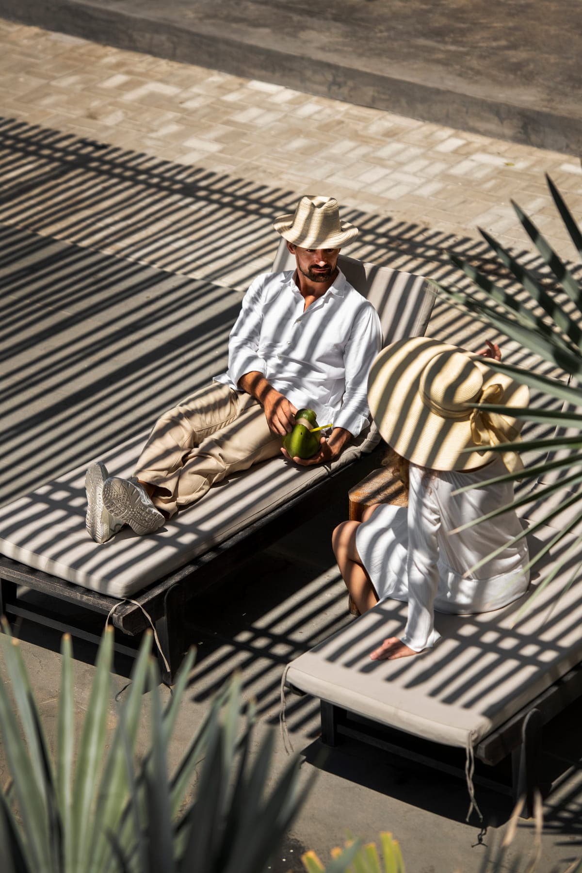 Two guests in white linen resting on striped daybeds under palm shadow at a resort poolside.