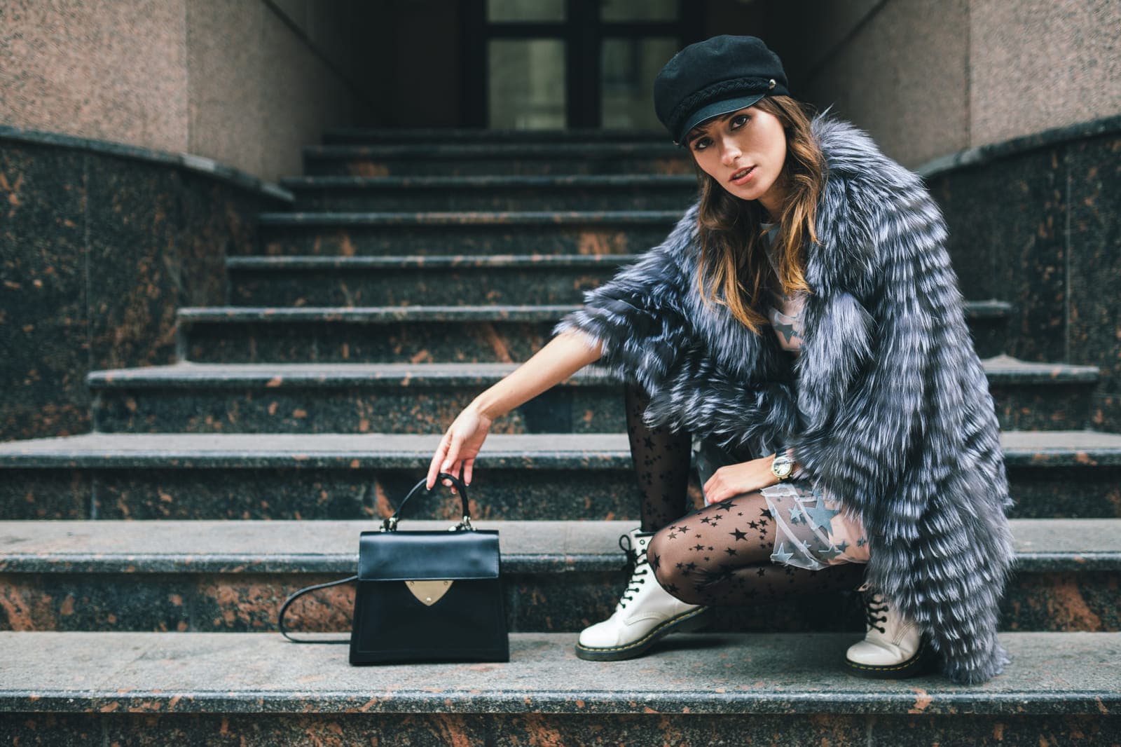 A woman in a silver fur coat seated on stone steps beside a black top-handle bag.