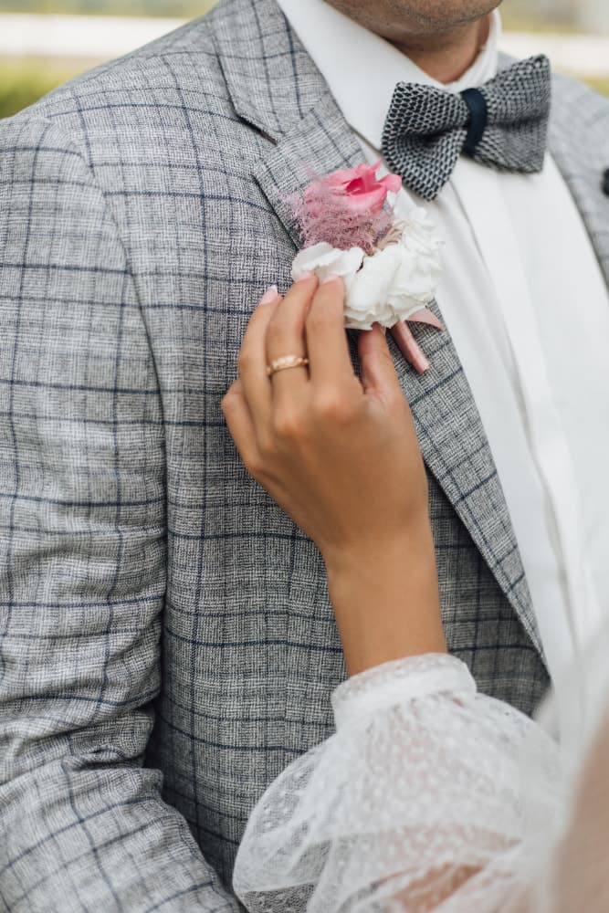 A hand pinning a pink and white boutonnière to the lapel of a grey checked suit jacket.
